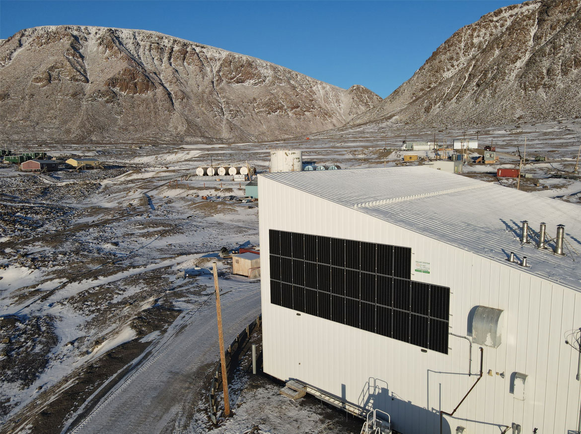 Plan large du centre communautaire avec les panneaux solaires sur sa façade et des réservoirs de diesel au loin, à côté des montagnes.