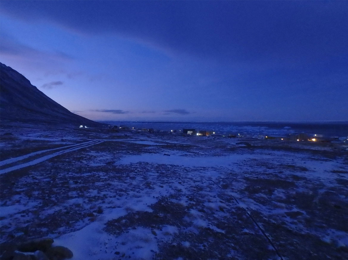 À gauche, le soleil se lève derrière une montagne et le hameau illuminé de Grise Fiord, au Nunavut, se trouve au loin.