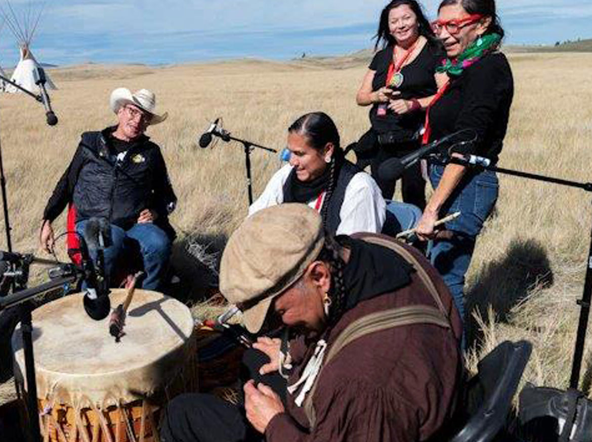 Cast and crew members participating in a drumming circle in a field.