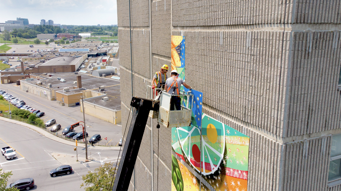 Men on a crane installing mural panels to the side of the building