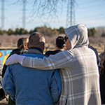 Deux personnes se tiennent à l'arrière d'un groupe, dos à la caméra. L'une des personnes a le bras autour de l'épaule de l'autre.