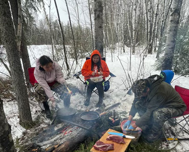 Three people sitting in folding chairs cooking food over a campfire.