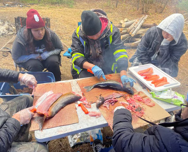 Group of youth seated around a table watching a man clean and prepare a fish.