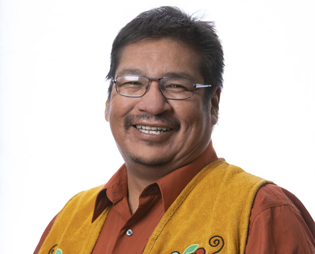 Chief Wayne Moonias against a white background wearing an orange collared shirt with a fringe vest that has beaded flowers on it.