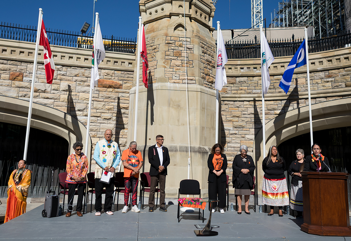 Des personnes debout sur une estrade à la Colline du Parlement lors de la cérémonie de lever du drapeau des survivants en 2025.