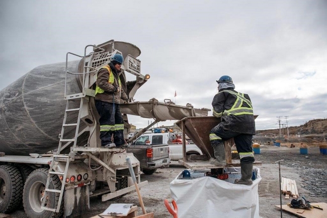 Un travailleur de la construction sur la plateforme d'un camion de ciment dirige le souffle du matériel de remblayage. Un autre travailleur sur une plateforme tient un grand entonnoir métallique servant à transporter le matériel dans le sous-sol. On voit des camions de travail en arrière plan.