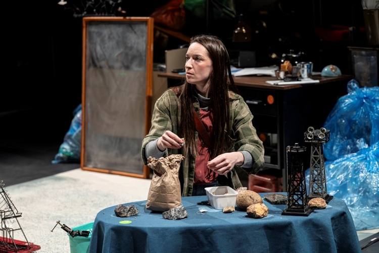 Une femme aux longs cheveux bruns, vêtue d'une chemise en flanelle, est assise à une table et examine des roches dans le cadre d'une pièce de théâtre.