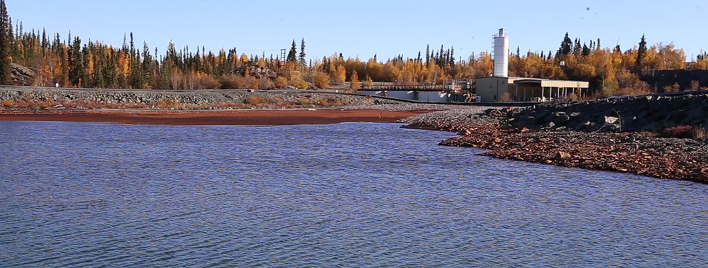 Cette photo montre la station d'épurement des effluents du site de la mine Giant à partir de l'autre côté d'un plan d'eau. La station comprend un ensemble de bâtiments et de grands bassins d'eau pour le traitement de l'eau. Des arbres se trouvent derrière le bâtiment et le long de la rive. On voit sur le rivage de l'eau de couleur rouille, en raison de son traitement à l'arsenic, séparée de l'eau propre que l'on voit à l'avant-plan.