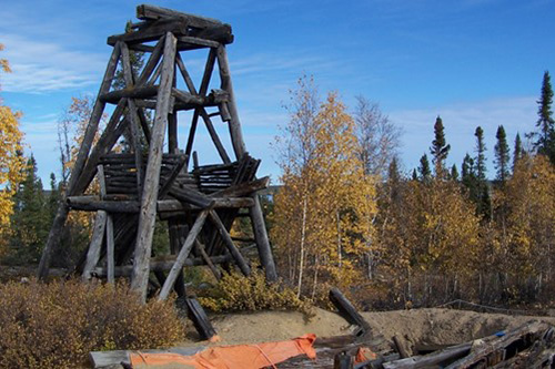 An old headframe made of wood stands above an open shaft surrounded by trees.