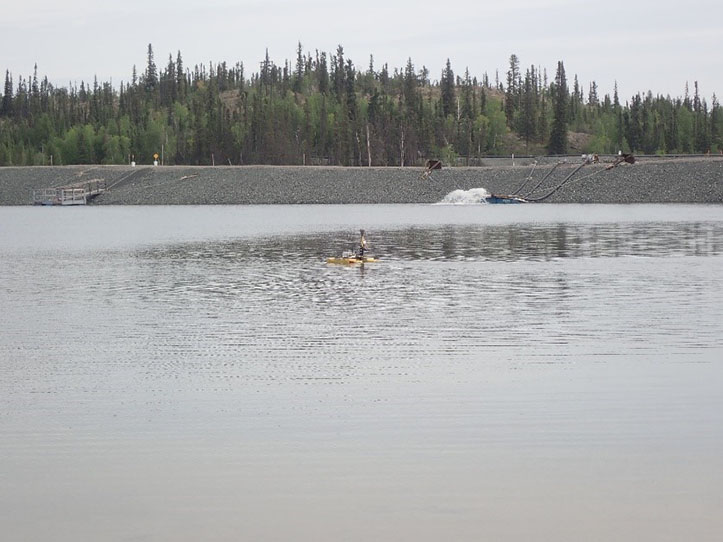This picture shows a hydro drone, which is a small remote controlled boat that monitors and maps depths of water, on the Polishing Pond on the Giant Mine site. In the background, trees are behind the rocky shore where pipes are discharging treated water into the pond