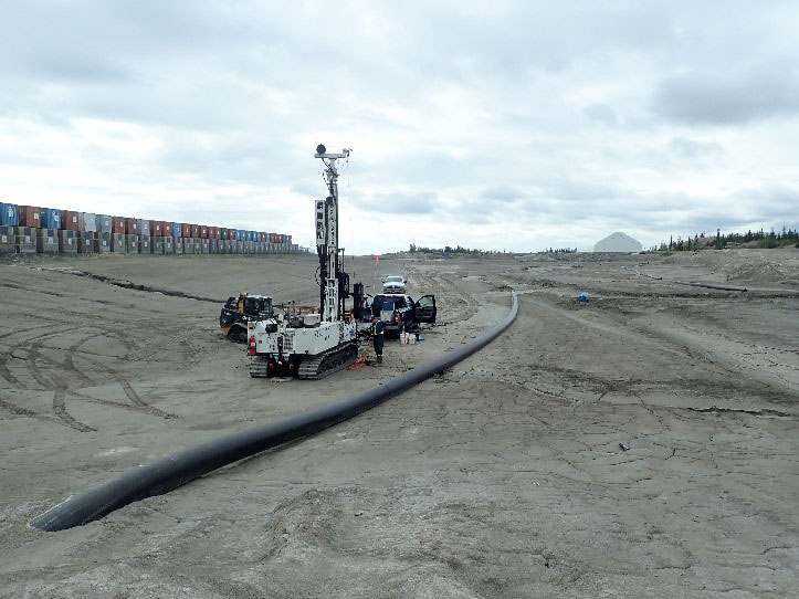 This picture shows a drilling rig, 2 trucks, and a Bobcat on a tailings pond, which is an expanse of grey processed rock. A worker in personal protective equipment overlooks the work. Sea cans stacked in the materials storage area are at the left of the photo, while a long black pipe which carries water from the south and central ponds to the North Pond runs beside the truck. Trees and a building are visible in the background.