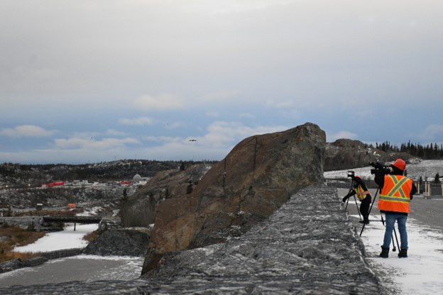 Deux journalistes vidéo équipés de caméras vidéo et vêtus de gilets de sécurité orange haute visibilité et de casques de sécurité enregistrent des images depuis une plate-forme rocheuse surplombant le site minier.