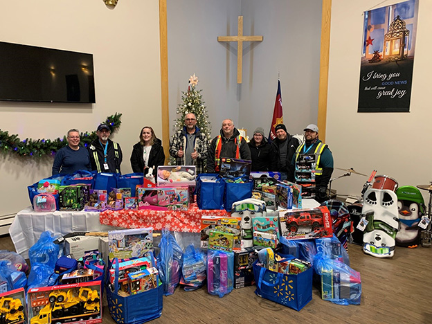 Un groupe de 8 adultes se tient derrière une table débordant de jouets non emballés pour une collecte de jouets de Noël.