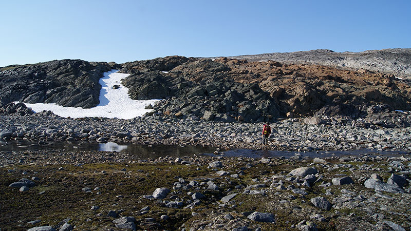 Barren-grounds tundra landscape with a geologist standing at the edge of a rocky creek, in front of a black and rusty brown bedrock outcrop
