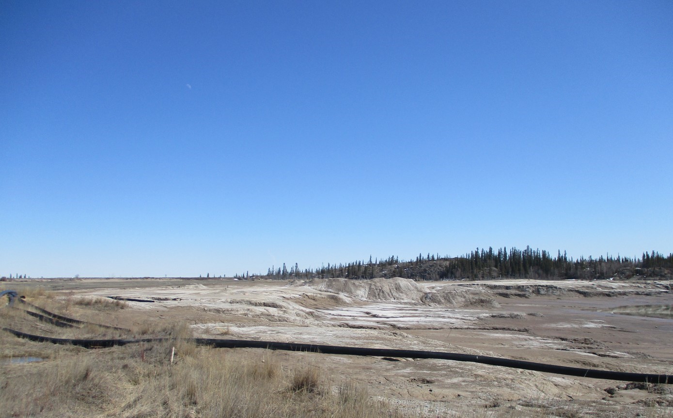 A tailings pond, where small pieces of rock and dust leftover from processing gold were deposited at the former mine sit, is shown. Some water is ponded in an area of the tailings. A rocky outcrop and trees are visible in the background and a pipe is visible in the foreground.