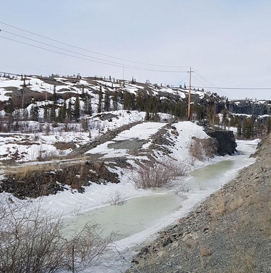 A frozen creek on the Giant Mine site showing signs of thawing on the surface. A rocky outcrop partially covered in snow is visible in the background.