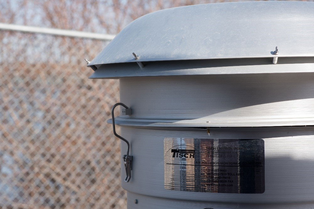 Cylindre métallique contenant de l'équipement de surveillance de la qualité de l'air, avec clôture à mailles losangées en arrière plan.