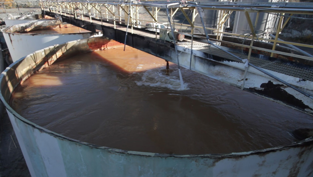 Cette photo montre une grande cuve d'eau traitée à l'usine saisonnière de traitement des effluents au site de la mine Giant. L'eau est d'une couleur rouge-brun en raison du sulfate ferrique utilisé pour que l'arsenic et d'autres métaux décantent dans l'eau. Une passerelle de métal est visible au-dessus de la grande cuve.