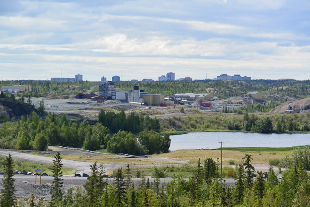 Cette photo montre une partie du site de la mine Giant en été. La route 4, ou route Ingraham, est à l'avant-plan, avec Baker Pond juste derrière. Les bâtiments du site sont visibles au milieu, et Yellowknife se trouve à l'horizon, à l'arrière-plan.