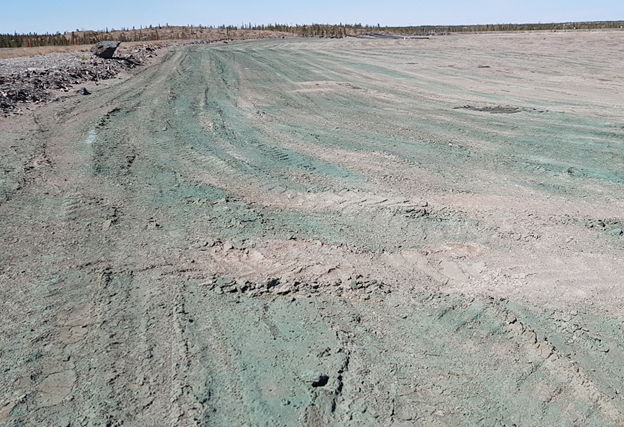 This image shows a dry tailings pond with trees and rocks in the far background. An acrylic co-polymer, made visible using a dye for the first application, was applied to form a crust that keeps the dust from becoming airborne.