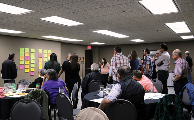 A group of people sitting at tables and standing in an open space in the room examine papers on a wall where they have written and categorized concerns about the Giant Mine site.