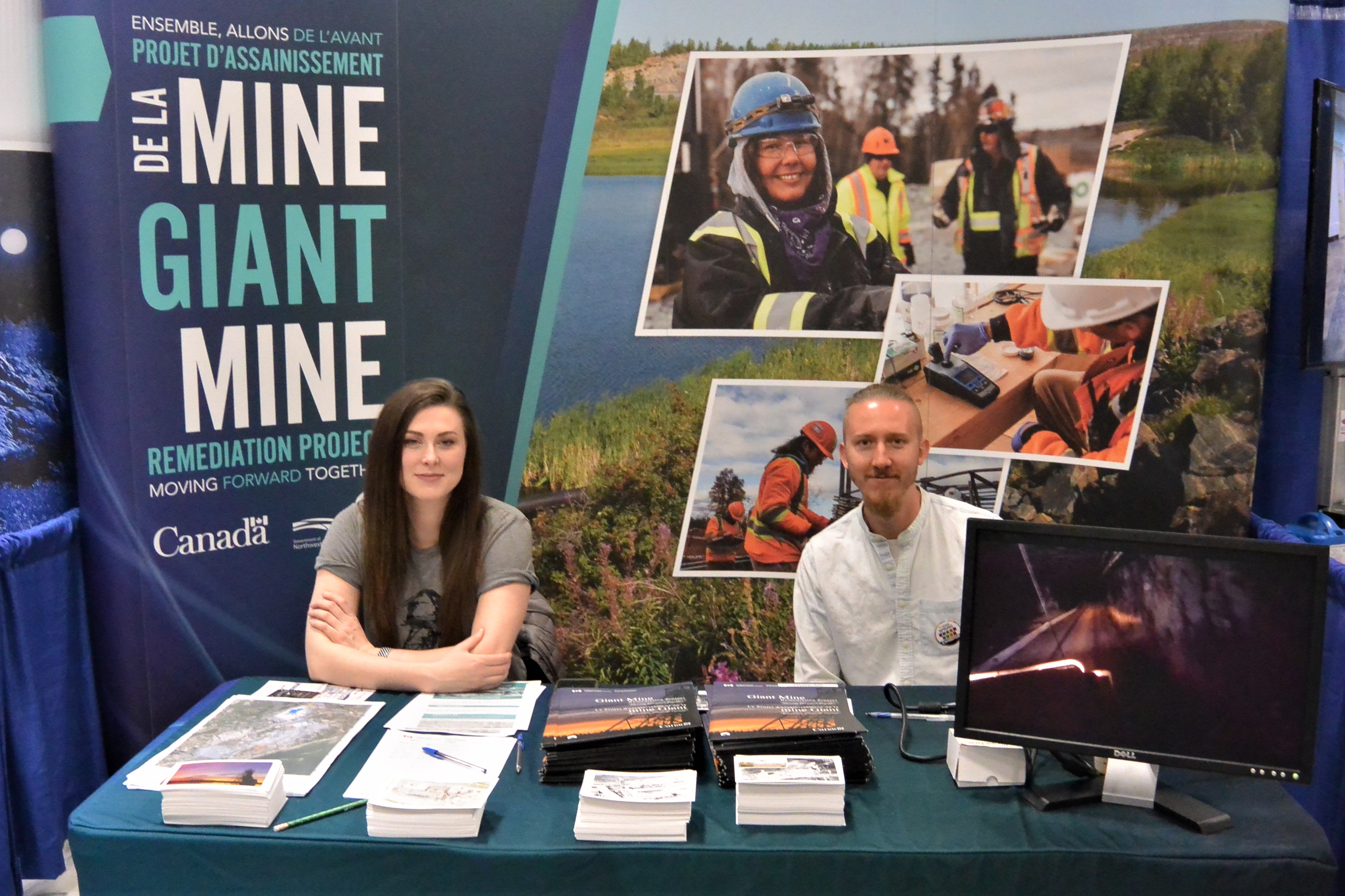 This pictures shows a woman and a man seated at a tradeshow booth table, with their Giant Mine Remediation Project backdrop behind them