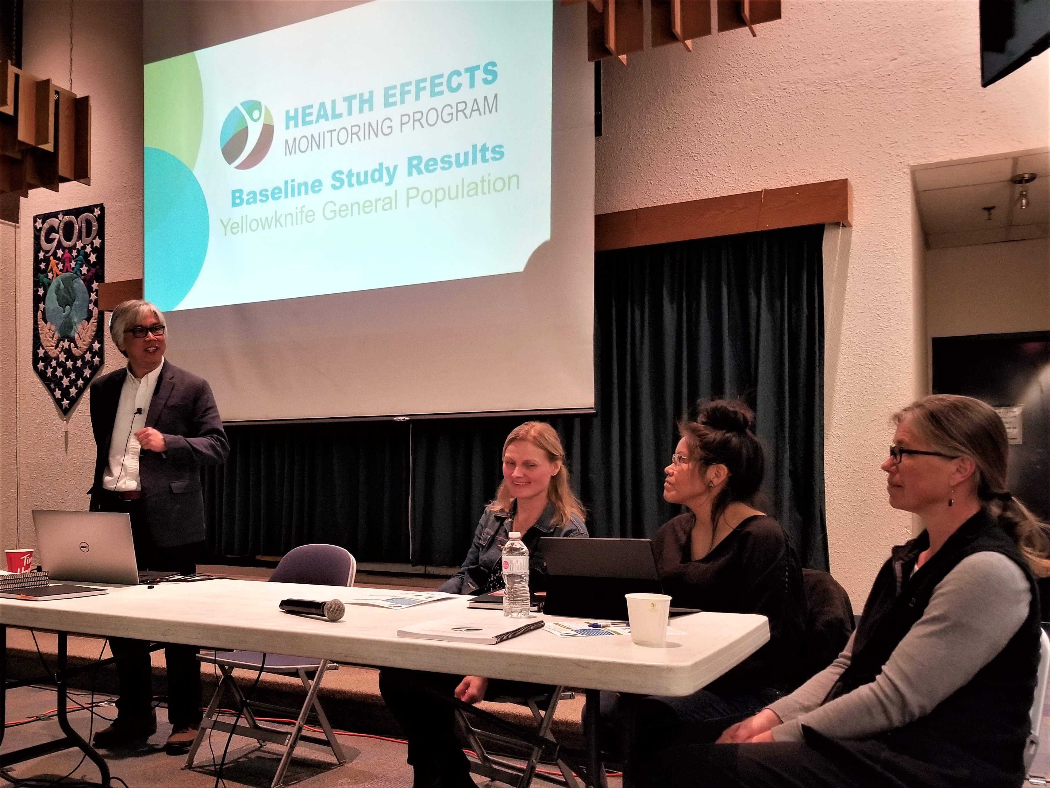 This picture shows a man standing behind a table doing a presentation. Seated to his right, at the table, are three women. Behind them, on a screen, a presentation slide reads 'Health Effects Monitoring Program, Baseline Study Results, Yellowknife General Population.'
