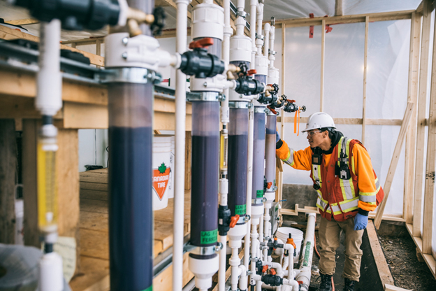 The picture shows a worker in PPE inspecting columns of adsorptive media that form a portion of the pilot water treatment plant at Giant Mine.