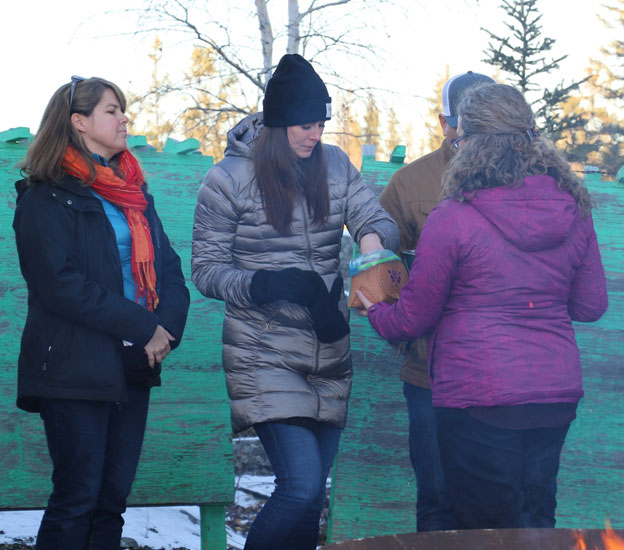This picture shows four people in winter clothing.  A woman reaches into a birch bark basket being held out by another woman.