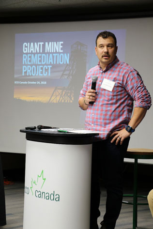 Un homme tenant un microphone est debout derrière un podium marqué du logo ECO Canada. Est affichée en arrière-plan une diapositive sur laquelle on peut lire « Giant Mine Remediation Project » (Projet d'assainissement de la mine Giant) et voir une photo du chevalement de l'ancien puits C au soleil couchant.