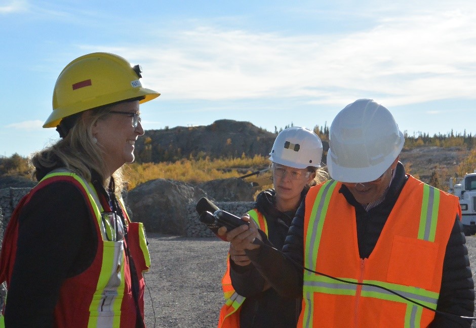 A man in a reflective vest and a hard hat holds a recording device in front of a smiling woman, also in personal protective equipment. Another woman in personal protective equipment listens in. A rocky outcrop, a flat gravel surface, and a work truck are partially visible in the background.