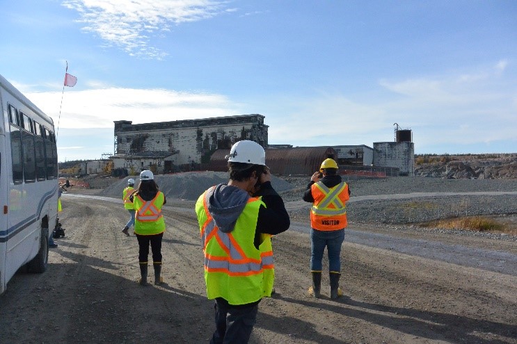 People in personal protective equipment stand on a gravel road, taking pictures of a dilapidated building. A bus is partially visible, parked on the road next to the people.