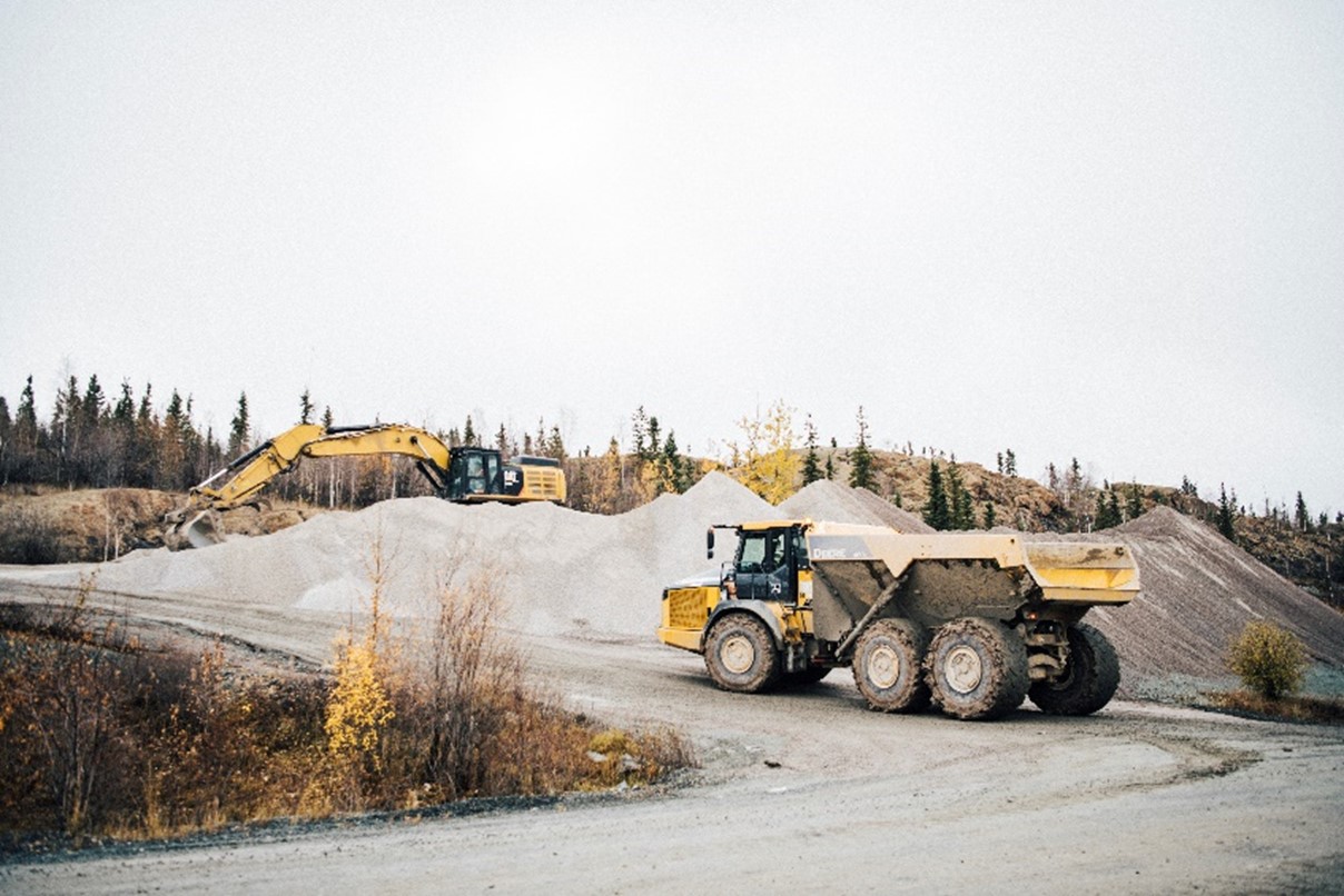 An empty dump truck drives on an unpaved road toward an excavator on a large pile of aggregate material