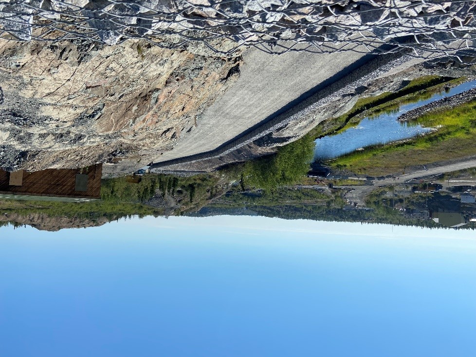 Une plateforme plate de gravier est visible à côté d'un affleurement rocheux. Une paroi rocheuse entourée d'un grillage fait office de barrière d'un côté. De vieux bâtiments, une route, un ruisseau et des arbres sont également visibles à côté de la plateforme.