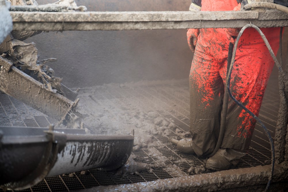 Du remblai en pâte cimentée est versé à travers une grille dans le sous-sol de la mine. Debout sur la grille, on voit un travailleur (vue partielle) portant un équipement de protection individuelle qui immobilise un tuyau avec sa botte.